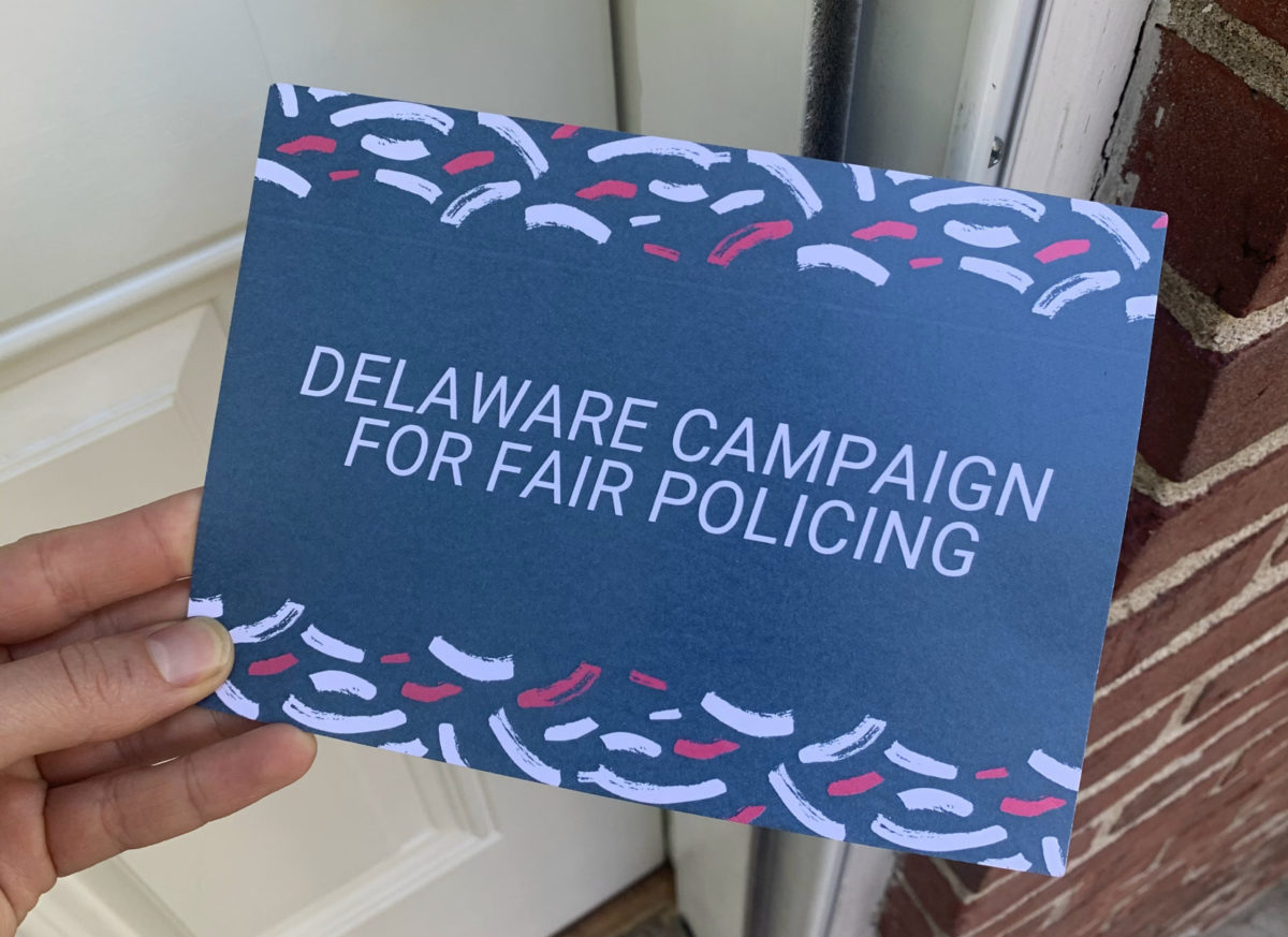 Hand holding a postcard that reads "Delaware campaign for fair policing" in front of a house door