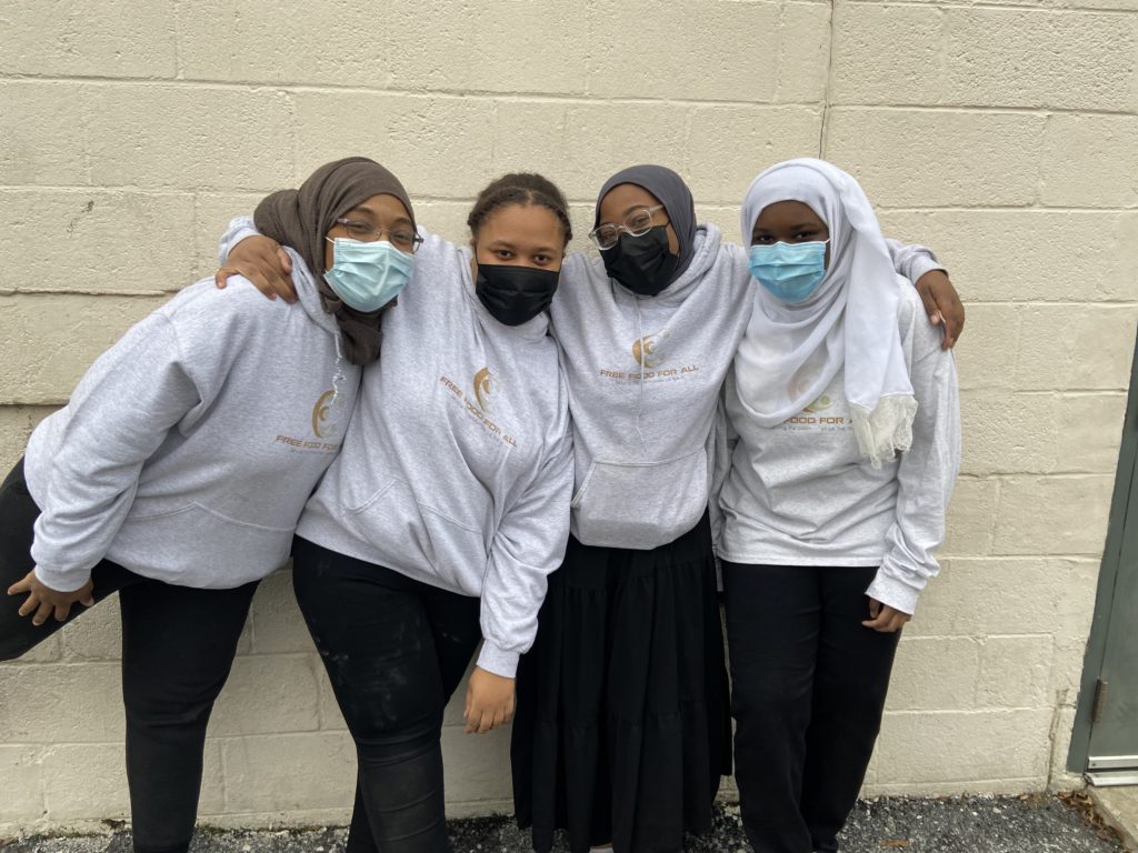 A group of four women in black pants and gray-white long-sleeve shirts, face masks, and head coverings; they have their arms around each other