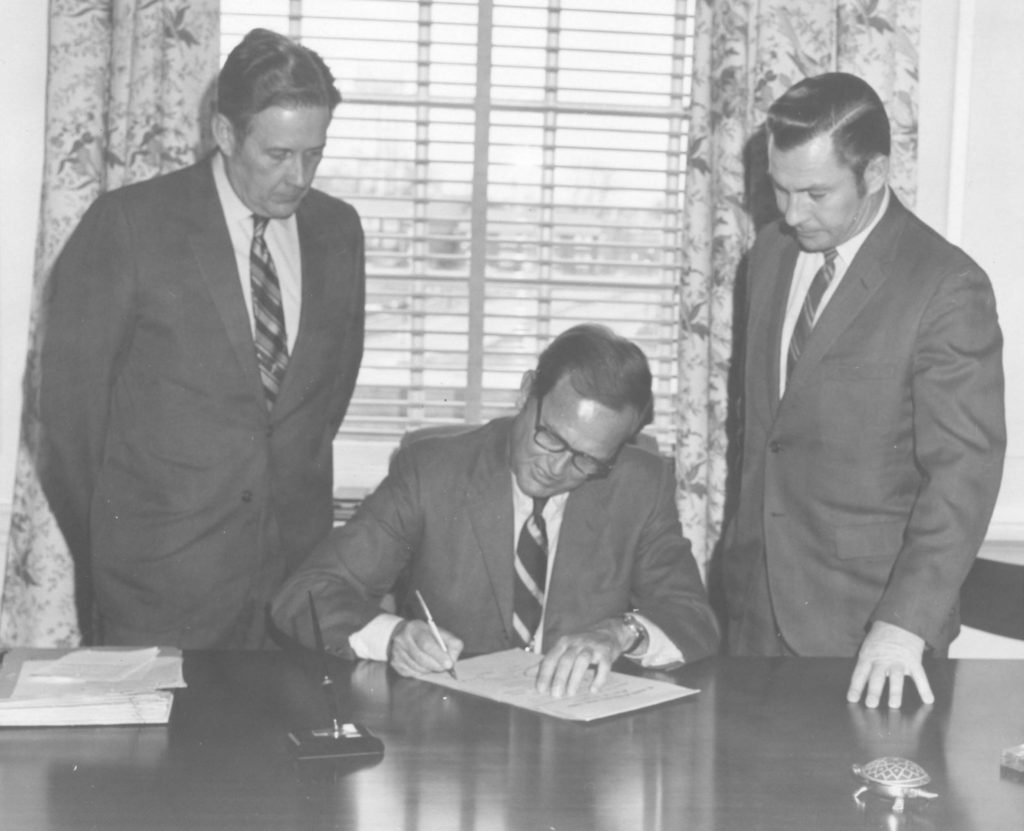 A man in the middle signing a paper at the desk, surrounded by two men standing above him